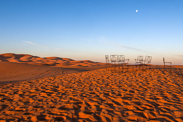 A tranquil Moroccan desert scene featuring windswept dunes and a delicate set of ornate metal chairs and a table under a clear blue sky, evoking solitude, travel, and outdoor dining.