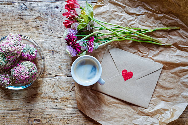 Cup of coffee, kraft envelope with a red heart, colorful bouquet and plate of sweets are arranged on crumpled brown paper over a rustic wooden table, evoking a slow, cozy morning for Valentine’s Day or Mother’s Day. The open wooden surface around the scene leaves plenty of copy space for promotional text, quotes, or greeting messages, while the overall mood feels comforting, loving, and relaxed.