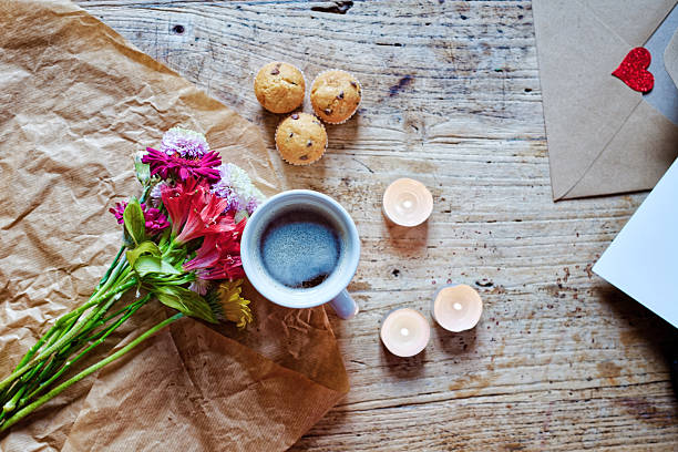 A steaming coffee, bright mixed flowers and glowing tea light candles are arranged on brown wrapping paper over a rustic wooden table, with mini cupcakes and an envelope just in frame, creating a romantic Valentine’s Day or Mother’s Day atmosphere with ample copy space for text or design elements.