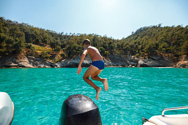 Child jumping into clear turquoise sea from a rented boat during summer family vacation near island coastline.