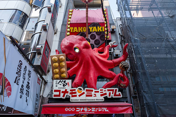 Osaka, Japan - November 5, 2023: A large red octopus sculpture holding takoyaki balls is mounted above the entrance of a restaurant in the Dotonbori district, with Japanese signage and neighboring buildings visible.