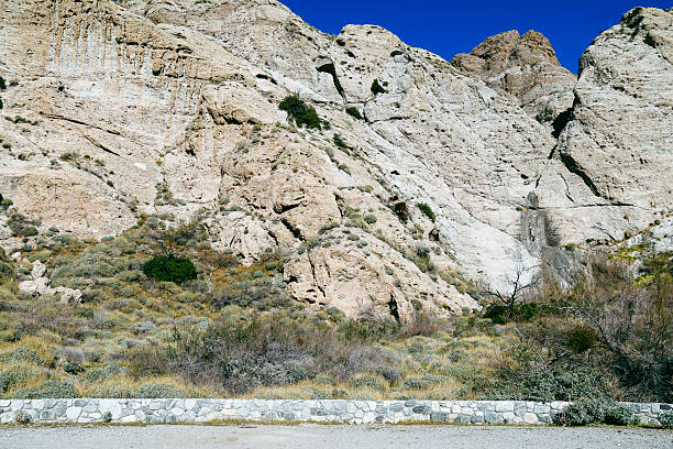 A rocky beige cliff rises above dry shrubs and sparse vegetation under a clear deep blue sky bordered by a low stone wall in a rugged desert mountain landscape
