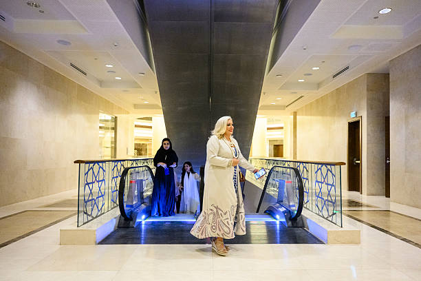 Three women in varying traditional and modern attire descending an escalator in a luxurious, contemporary shopping mall with intricate Arabian architectural details