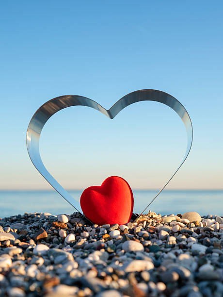 A romantic composition featuring a soft red heart placed inside a metallic heart-shaped cutter on a pebble shore.