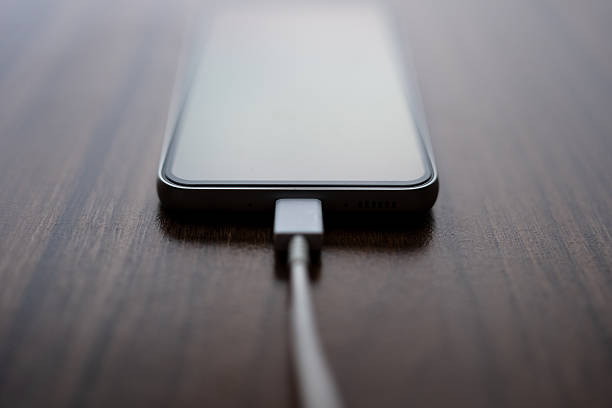 Minimal close-up of a modern smartphone plugged into a charging cable on a wooden desk. Clean and elegant composition representing mobile technology, power supply, battery charging, connectivity, and everyday digital life. Shallow depth of field with copy space.