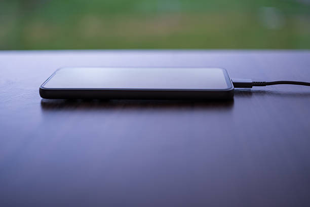 Modern smartphone lying on a wooden table and connected to a charging cable. Clean and minimal composition with blurred green background, representing mobile technology, battery life, energy, power supply, and everyday digital connectivity. Ideal for technology and lifestyle concepts.