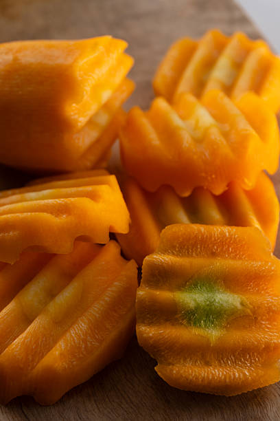 A close-up, high-angle shot captures a pile of fresh, crinkle-cut carrot slices resting on a textured wooden board. The vibrant orange vegetable pieces exhibit a distinct wavy shape created by a crinkle cutter, with a moist and shiny surface that reflects the light. In the foreground, one slice displays a faint green core, adding color variation to the monochromatic orange palette. The background is a soft, out-of-focus wooden tone, emphasizing the sharp detail and texture of the raw ingredients prepared for cooking or snacking.
