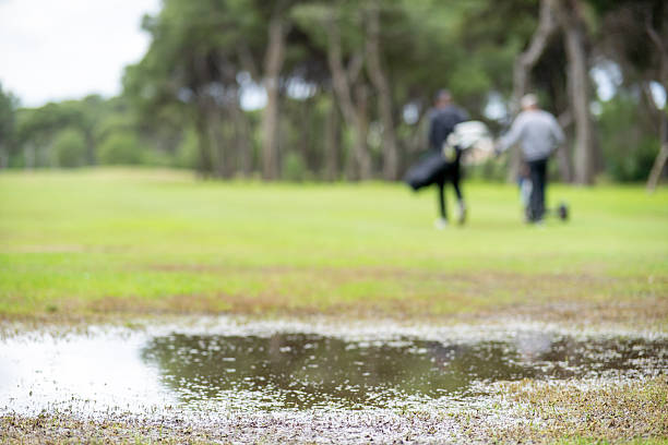 A man and a woman are walking on a golf course. The woman is holding a golf bag and the man is holding a golf club. The background is a wet, muddy area