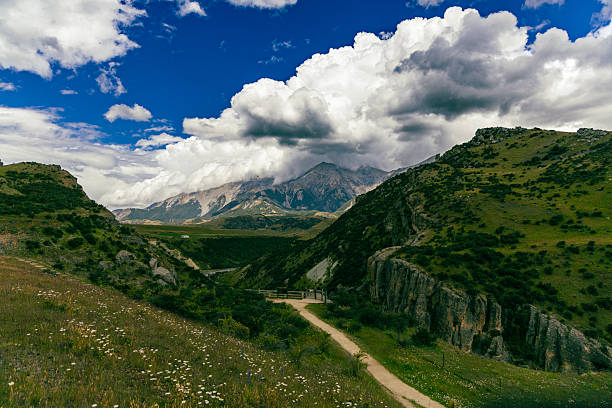 Remote footpath winds through lush hills beneath towering peaks cloudscape