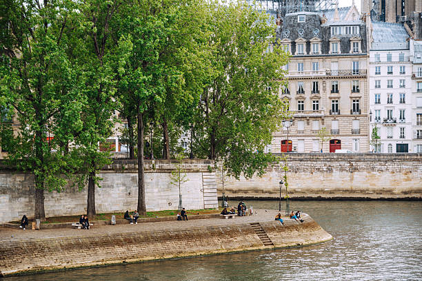 Lush trees line a stone quay along the Seine in Paris. People relax by the water while classic Parisian buildings rise in the background, creating a calm urban riverside scene.