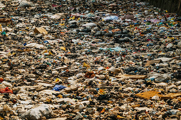 A crowded Mumbai slum line along a polluted canal, with makeshift houses, scattered trash, and overgrown edges. The image conveys urban poverty, neglect, and environmental decay in a dense city corner.