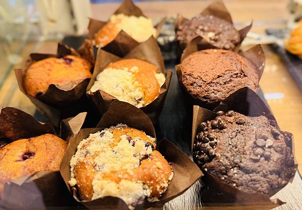Assorted Fresh Muffins On Bakery Display, Pastry Selection Presented In Tulip Liners With Crumb, Streusel And Chocolate Toppings, Warm Golden Lighting Creating Cozy Cafe Atmosphere, Artisan