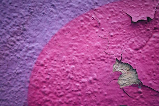 Close-up view of textured building wall with peeling paint. The surface features vibrant pink and purple colors, revealing a rough, damaged area underneath