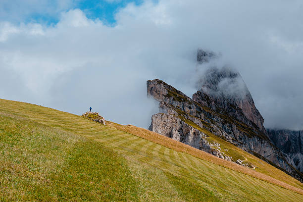 A solitary figure stands on a green mountain pasture, gazing at the imposing Seceda ridgeline partially obscured by atmospheric fog