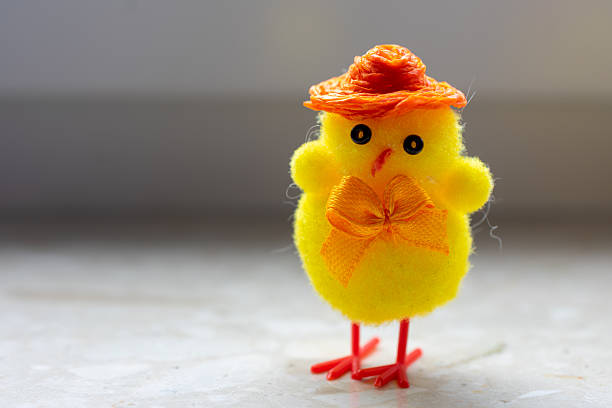 Yellow stuffed chicken with a hat and a bow. It is standing on a white surface. The hat is orange and the bow is orange.