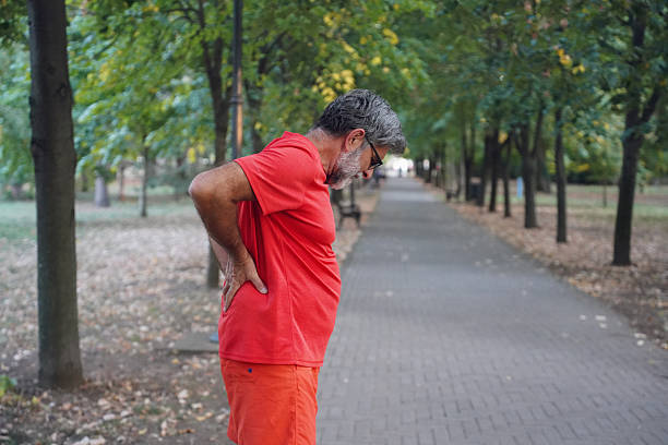 Senior man exercising outdoors in a city park in the morning. Older adult performing stretching and warm-up exercises with visible physical discomfort while maintaining an active routine. Concept of aging, mobility challenges, body awareness, fitness routine, active lifestyle, outdoor exercise, realistic senior fitness, and everyday wellness in later life.