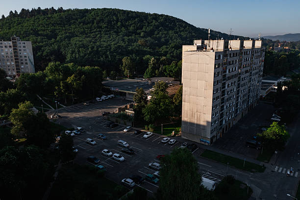 A high-rise residential building stands beside a parking lot and a lush, forest-covered hill, illuminated by low-angle sunlight. The long shadows and deep contrasts evoke a quiet, end-of-day atmosphere in this suburban setting. The image captures the juxtaposition of urban infrastructure and natural landscape.