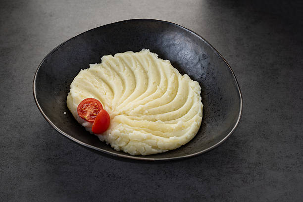 Creamy mashed potatoes served in a dark bowl with a cherry tomato slice. Mashed potatoes are artistically arranged in a black bowl with a cherry tomato on the side.