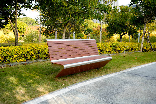 A modern, minimalist park bench made of wood and metal, placed on grass near a paved pathway in a well-designed urban park. The surrounding area features neatly trimmed hedges, trees with autumn foliage, and soft natural lighting. Ideal for themes of public space, urban design, sustainability, and peaceful outdoor environments.