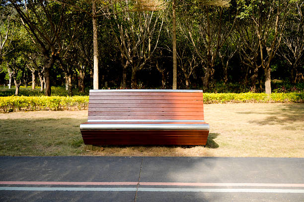 A modern, minimalist park bench made of wood and metal, placed on grass near a paved pathway in a well-designed urban park. The surrounding area features neatly trimmed hedges, trees with autumn foliage, and soft natural lighting. Ideal for themes of public space, urban design, sustainability, and peaceful outdoor environments.