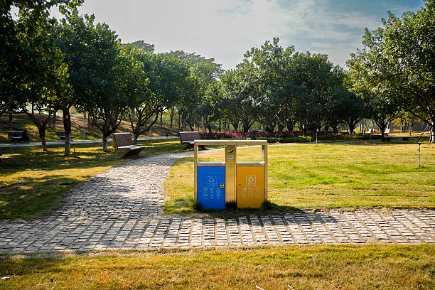 A pair of color-coded recycling bins — blue and yellow — stand on a paved pathway in a spacious urban park. The bins are clearly marked for different types of waste, promoting proper disposal and environmental awareness. Surrounding the area are lush green lawns, mature trees providing shade, and wooden benches in the distance. Bright sunlight enhances the natural beauty of the scene. Ideal for themes of sustainability, public cleanliness, urban landscaping, and eco-conscious city living.\