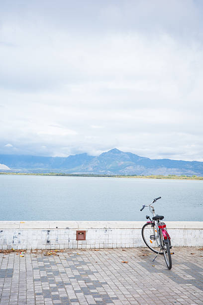 A single bicycle resting against a low wall along a quiet lakeside promenade, overlooking calm water and distant mountains beneath a soft cloudy sky. The minimal composition and generous copy space evoke freedom, solitude, slow travel, and mindful living. Ideal for themes of lifestyle, sustainable transport, urban nature, travel inspiration, and peaceful moments