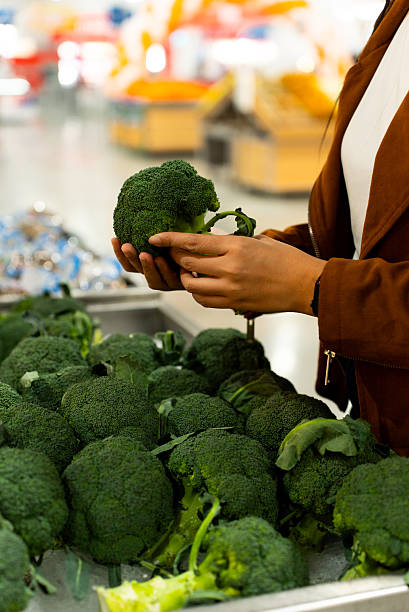 Vertical woman's hand holding broccoli fresh vegetable while shopping at grocery store for healthy lifestyle, female looking for fresh food and daily household essential in modern supermarket.