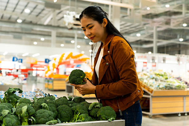 Young asian woman holding broccoli fresh vegetable while shopping at grocery store for healthy lifestyle, female looking for fresh food and daily household essential in modern supermarket.