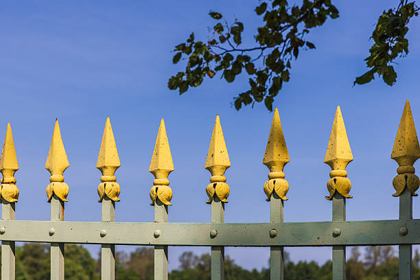 A decorative yellow spiked fence line under a bright blue sky. The weathered metal posts and ornamental spikes create a classic garden perimeter barrier, with leafy branches framing the scene