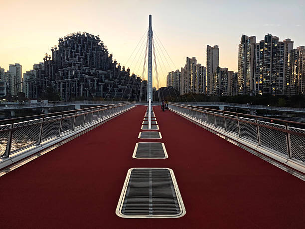 People walking at sunset on the Baihe Bridge, a recently opened modern suspension pedestrian bridge over Suzhou Creek, connecting Jing'an and Putuo districts, Shanghai, China. In the distance, the Tian An 1000 Trees, a mixed-use development designed by Heatherwick Studio that features a unique architecture with over 1,000 trees and 250,000 plants covering its \