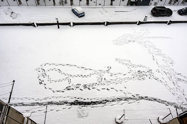 Top-down view of fresh snow covering a ground surface, marked with numerous human footprints and car tracks. In the background, parked cars are partially visible near a fence.