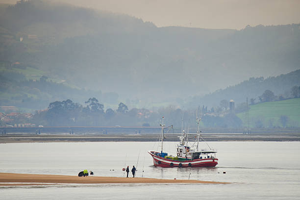 Fishing boat passing people fishing on a sandbar with misty mountains and a coastal landscape in the background. Santoña, Cantabria