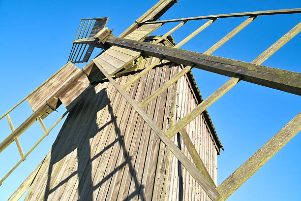 Historic wooden windmill on a rural meadow under a blue sky with white clouds, surrounded by fields and trees, creating an idyllic, tranquil landscape atmosphere.