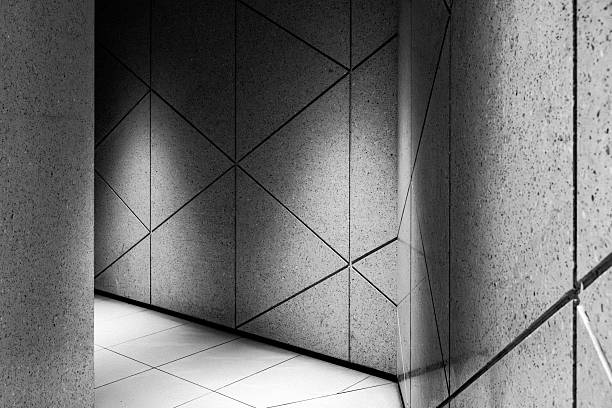This monochromatic image captures a sleek architectural detail, likely within a contemporary corridor or lobby. The focus is on the wall design, which features large panels divided by deep grooves into a striking pattern of intersecting triangles. The surface has a fine, speckled texture resembling concrete or stone, which is highlighted by soft, dramatic lighting that creates subtle gradients of shadow. A polished, white tiled floor is visible at the base, contrasting with the textured walls and enhancing the clean, brutalist-inspired aesthetic.