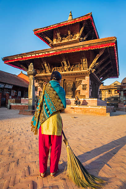 Nepali young woman sweeping on a d Durbar Square in Bhaktapur. An ancient Hindu temple on the background. Bhaktapur is an ancient town in the Kathmandu Valley and is listed as a World Heritage Site by UNESCO for its rich culture, temples, and wood, metal and stone artwork.