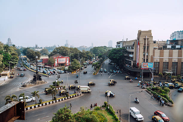 A vibrant street scene in Mumbai shows a busy intersection with cars, buses, and motorcycles, flanked by palm trees and mid-rise buildings. The image captures urban life, traffic flow, and city bustle.