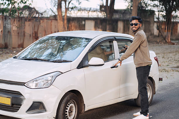Young man wearing sunglasses standing beside a white car and holding a smartphone on a city street. Concept of smart mobility, urban transportation, and modern lifestyle.