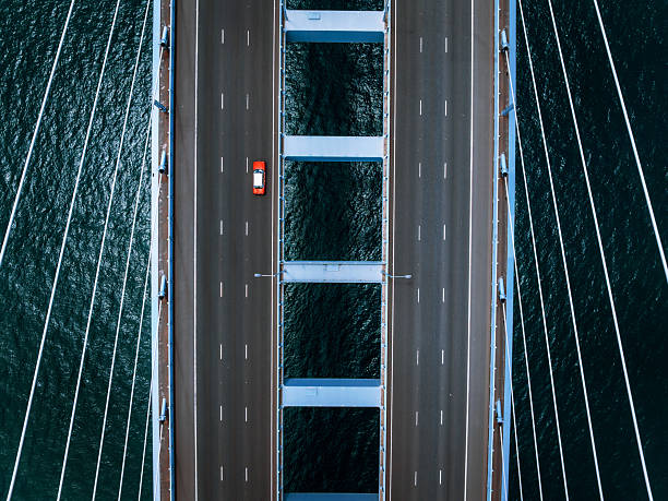 Aerial drone shot of a large suspension bridge with multiple lanes full of cars, spanning over blue-green water. Symmetrical cables frame the road deck, emphasizing engineering and travel.