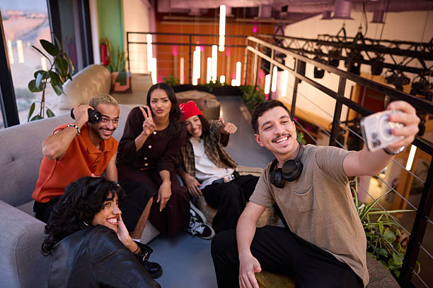 Group of friends spending leisure time at modern indoor space. Smiling man is taking selfie with buddies. They are sitting on couch.