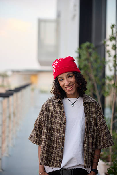 Portrait of smiling young female in casual clothes posing on terrace. Adult female wearing fully unbuttoned shirt along with red beanie. She is looking at camera.