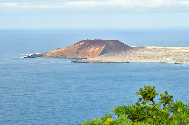 A view of a volcanic island with a prominent red cone surrounded by the blue ocean. Several sailboats are anchored near the sandy shore under a cloudy sky.
