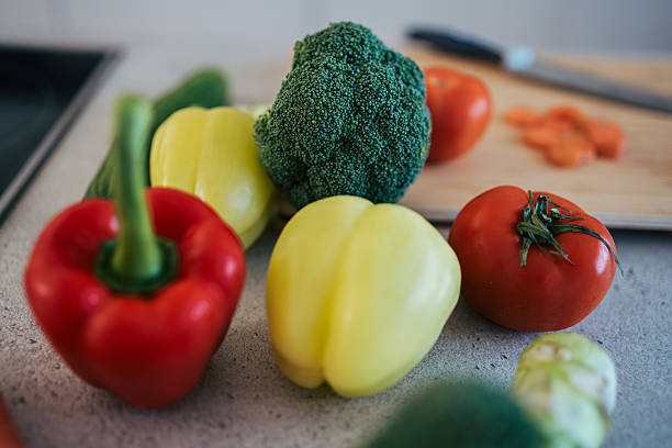 Fresh red and yellow bell peppers, broccoli, and tomatoes sitting on a kitchen counter with a cutting board and knife in the background, preparing a healthy meal