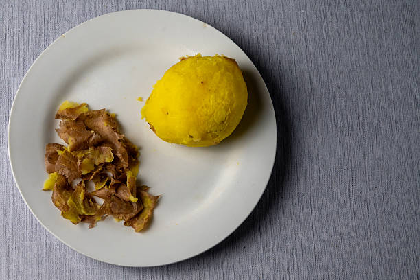 Top view of a single boiled potato on a white plate with potato peel, symbolizing food poverty, rising living costs, minimal nutrition, and economic hardship.