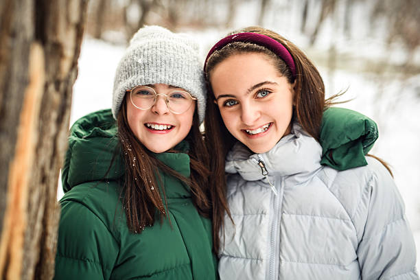 Two happy young women are embracing and smiling in a winter forest, wearing warm winter clothes