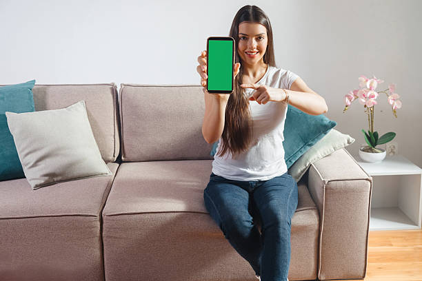 A woman with long hair sits on a couch showing a mobile device with a blank green screen. She smiles and points at the device in a living room with modern decor.
