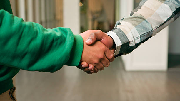 Two people shake hands in a bright indoor space, symbolizing partnership, trust, and collaboration. A green sleeve and plaid shirt highlight diverse professionals sealing a deal or agreeing on a plan.