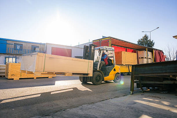 Worker driving forklift loading shipment carton boxes goods on wooden pallet at loading dock from container truck to warehouse cargo storage in freight logistics, transportation industrial, delivery