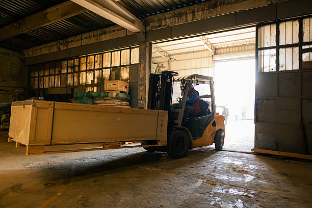 Worker driving forklift loading shipment carton boxes goods on wooden pallet at loading dock from container truck to warehouse cargo storage in freight logistics, transportation industrial, delivery