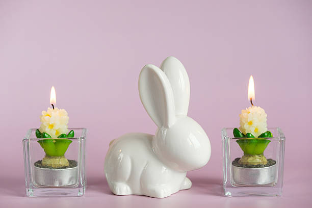 Easter still life with a white ceramic rabbit and two burning candles in glass candlesticks on a pink background. The concept of comfort, celebration and bright Easter.