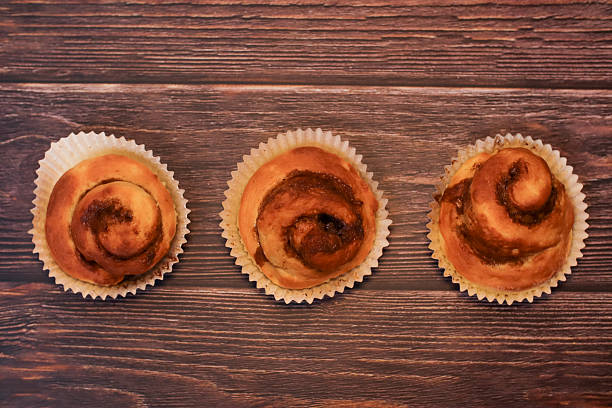 Three freshly baked cinnamon swirl sweet buns presented in paper liners on a dark, rustic wooden table background.